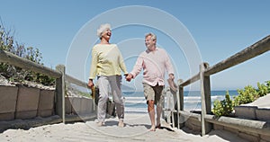 Happy senior caucasian couple holding shoes in their hands walking on path leading to the beach