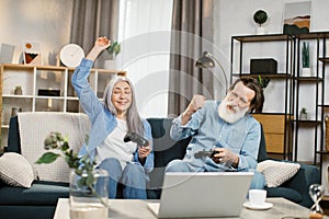 Happy senior caucasian couple sitting on couch with wireless joysticks in hands