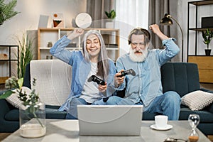 Happy senior caucasian couple sitting on couch with wireless joysticks in hands