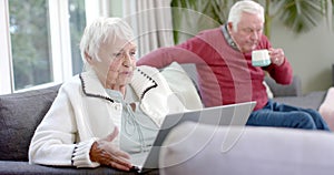 Happy senior caucasian couple sitting on sofa using laptop for video call at home, slow motion