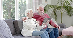 Happy senior caucasian couple sitting on sofa using tablet at home, slow motion