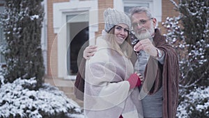 Happy senior Caucasian man hugging young wife and bragging keys from new house at camera. Smiling grey-haired