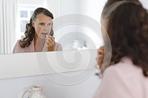 Happy senior caucasian woman in bathroom, looking in mirror, applying lipstick