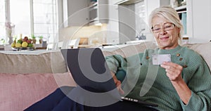 Happy senior caucasian woman sitting on sofa in living room, using laptop and holding credit card