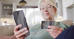 Happy senior caucasian woman on sofa in living room, using smartphone and holding credit card