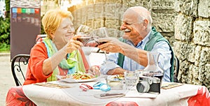 Happy senior couple cheering with red wine in italian restaurant outdoor - Old people having fun in vacation holidays eating lunch