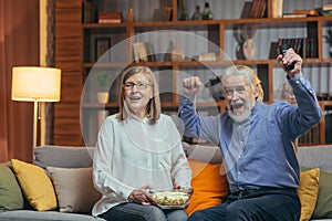 Happy senior couple cheers in watching sport competitions television TV game celebrating victory together on sofa in living room