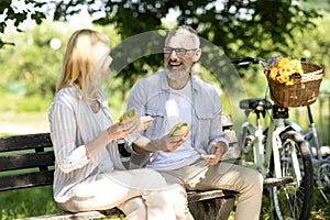 Happy Senior Couple Eating Sandwiches While Sitting On Bench In Park