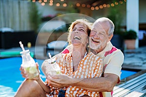 Happy senior couple enjoying drinks when relaxing and sitting by swimming pool in summer.