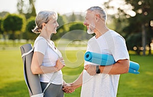 Happy senior couple with exercise rubber mats holding hands smiling and standing on park lawn