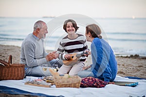 Happy senior couple with granddaughter sitting on blanket and having picnic outdoors on beach by sea.