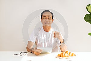 Happy senior couple having croissant breakfast at home and drinking milk, touching on digital tablet