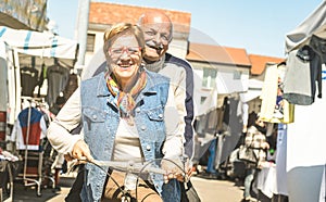 Happy senior couple having fun on bicycle at city market - Active playful elderly concept riding bike at retirement time