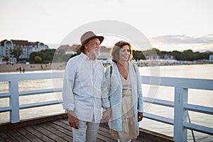 Happy senior couple hodling hands on walk outdoors on pier by sea, looking at view.