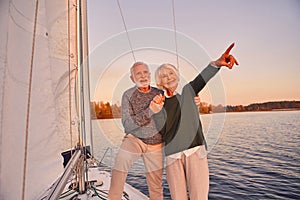 Happy senior couple holding hands and smiling while standing on the side of yacht deck floating in sea, woman pointing