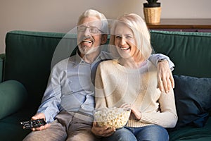 Happy senior couple holding remote control snack laughing watching tv