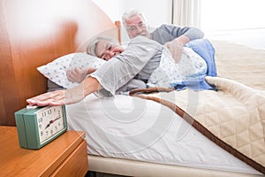 Happy senior couple at home stop alarm in morning wakeup - woman touch colck to continue sleeping man hugging her - window light