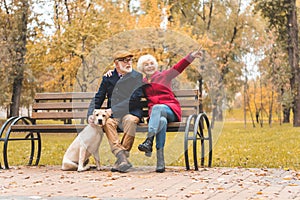 happy senior couple with labrador retriever dog sitting on bench in autumn