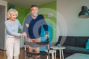 Happy senior couple living together in nursing home, woman helping her husband to walk inside the apartment