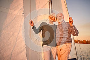 Happy senior couple in love hugging and enjoying amazing sunset while standing on the side of sailboat or yacht deck