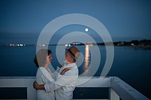 Happy senior couple in love hugging outdoors on pier by sea at moonlight, looking at each other.