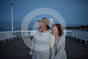 Happy senior couple in love on walk holding hands outdoors on pier by sea at moonlight, looking at view.