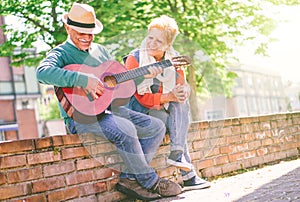 Happy senior couple playing a guitar while sitting outside on a wall on a sunny day