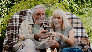 Happy Senior Couple Relaxing On Chairs In Garden And Using Smartphone