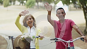 Happy senior couple sitting on bikes and waving hands at camera