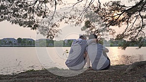 Happy senior couple sitting near lake view hugging at tree in evening sunset. smiley sweet asian elderly lover outdoor romantic sh