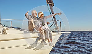 A happy senior couple sitting on the side of a sail boat on a calm blue sea, pointing at landscape, enjoying view