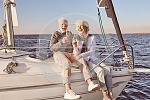 Happy senior couple sitting on the side of sail boat or yacht deck floating in sea. Man and woman drinking wine or