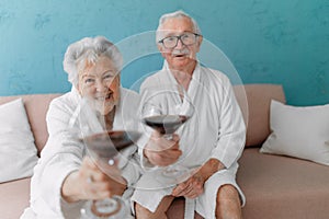 Happy senior couple sitting together in bathrobe on sofa with glass of wine, having nice time at home.
