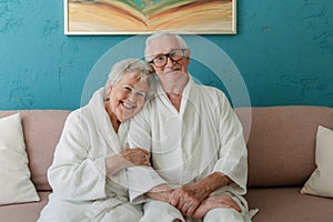 Happy senior couple sitting together in bathrobe on sofa, having nice time at home.