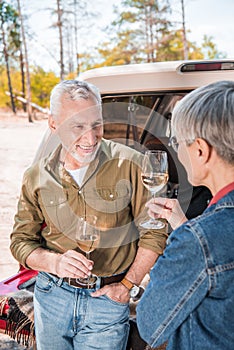 happy senior couple standing near car with wine glasses and looking at each other.