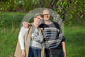 Happy senior couple tourists walking in green spring city park. Smiling elderly married in glasses traveling together.