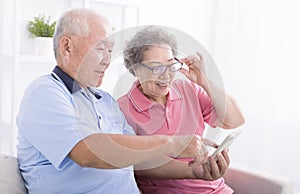 Happy senior couple using cell phone together at home, surfing internet, sitting on couch