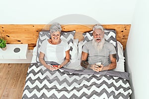 Happy senior couple using mobile smartphones below blankets in bed - Mature people having funny bed time