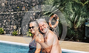 Happy senior couple on vacation in swimwear sitting at the swimming pool - man with beard and white hair hugs his wife laughing.