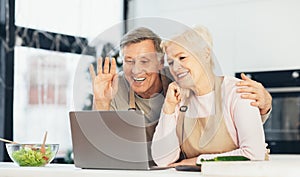 Happy Senior Couple Video Calling Using Laptop Sitting In Kitchen