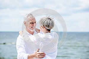 happy senior couple in white shirts embracing and looking at each other under blue sky.