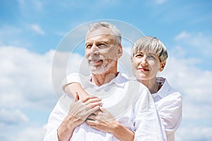 happy senior couple in white shirts embracing under blue sky.