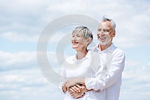 happy senior couple in white shirts embracing under blue sky.