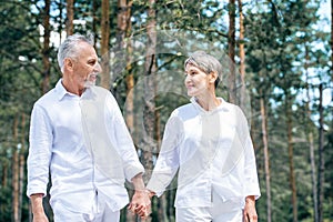 happy senior couple in white shirts holding hands and looking at each other in forest.