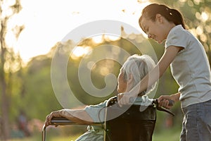 Happy senior disabled woman in wheelchair with granddaughter walking outside at park on sunny day,smiling child girl and elderly