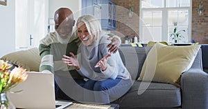 Happy senior diverse couple in living room sitting on sofa, using laptop, making video call