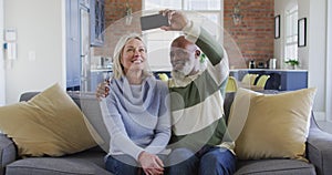 Happy senior diverse couple in living room sitting on sofa, using smartphone, making video call