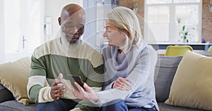 Happy senior diverse couple in living room sitting on sofa, using smartphone, making video call