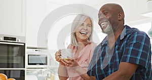 Happy senior diverse couple sitting in living room and drinking coffee