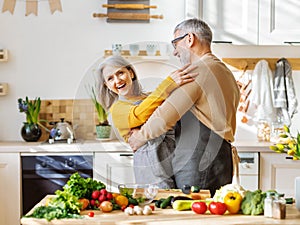 Happy senior elderly couple husband and wife embracing and dancing while cooking together in kitchen